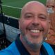 Homem careca sorrindo em primeiro plano com equipamento de TV e campo de futebol ao fundo