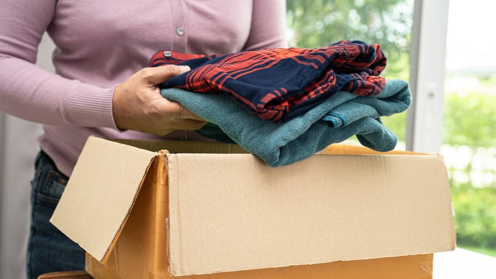 Mãos de pessoa adulta seguram roupas dobradas (camisa xadrez, peça azul) sobre caixa de papelão [Créditos: depositphotos.com / chormail@hotmail.com]