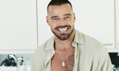 Cantor Ricky Martin sorrindo, cabelo e barba escura, camisa clara aberta, colar com dog tag