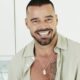 Cantor Ricky Martin sorrindo, cabelo e barba escura, camisa clara aberta, colar com dog tag