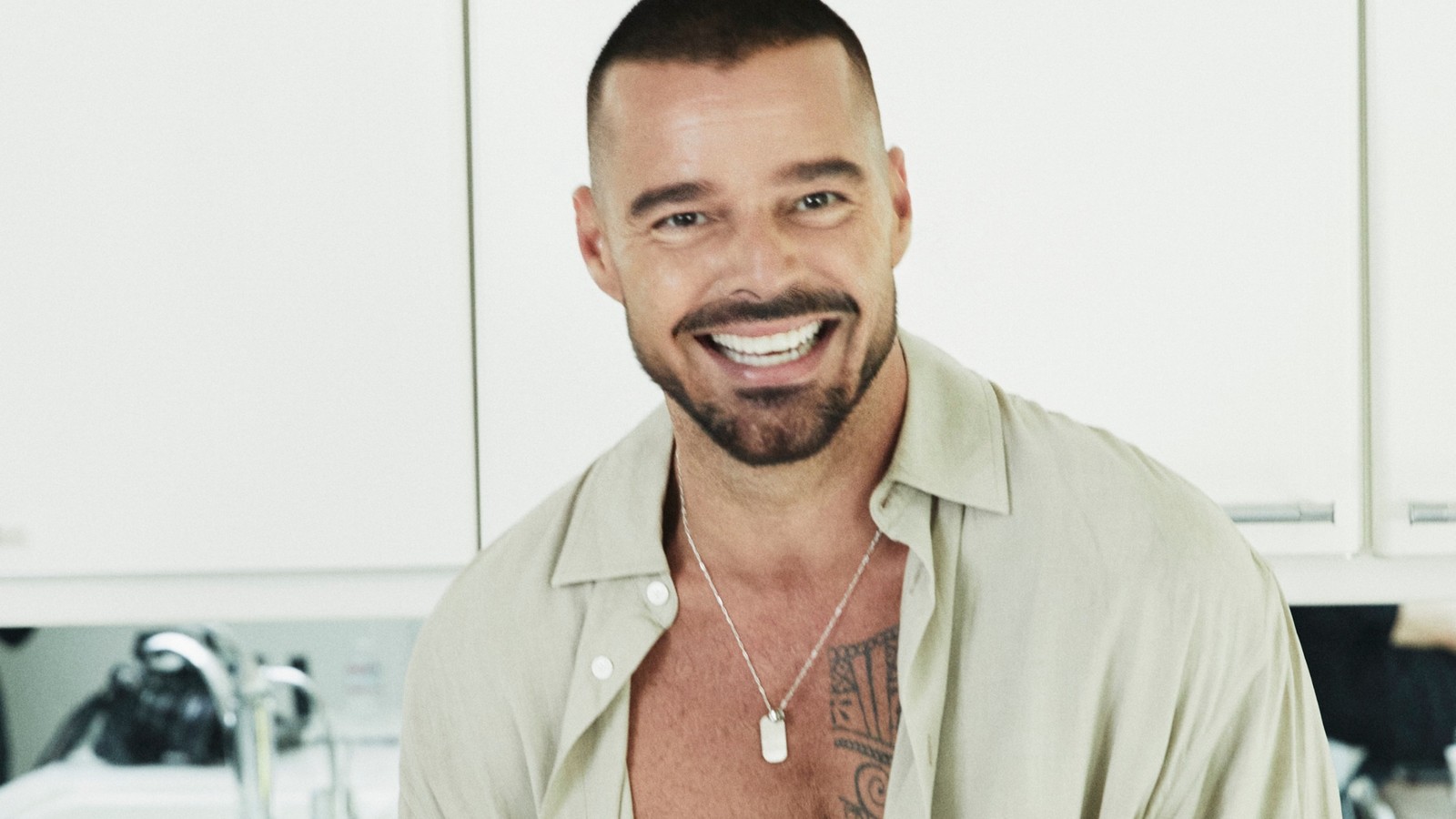 Cantor Ricky Martin sorrindo, cabelo e barba escura, camisa clara aberta, colar com dog tag