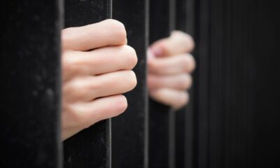 Duas mãos segurando barras de prisão pretas, com a mão direita desfocada ao fundo