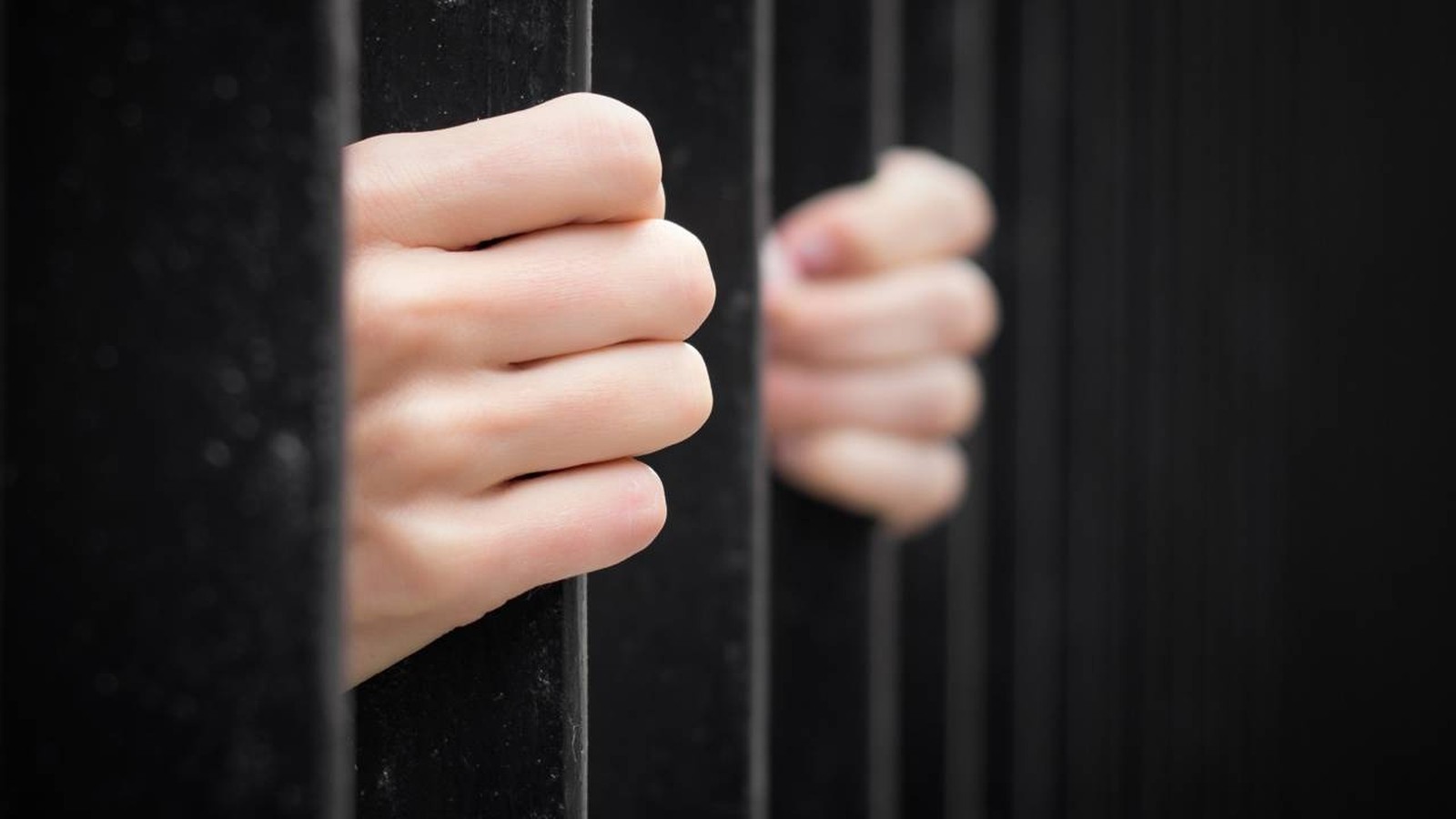 Duas mãos segurando barras de prisão pretas, com a mão direita desfocada ao fundo