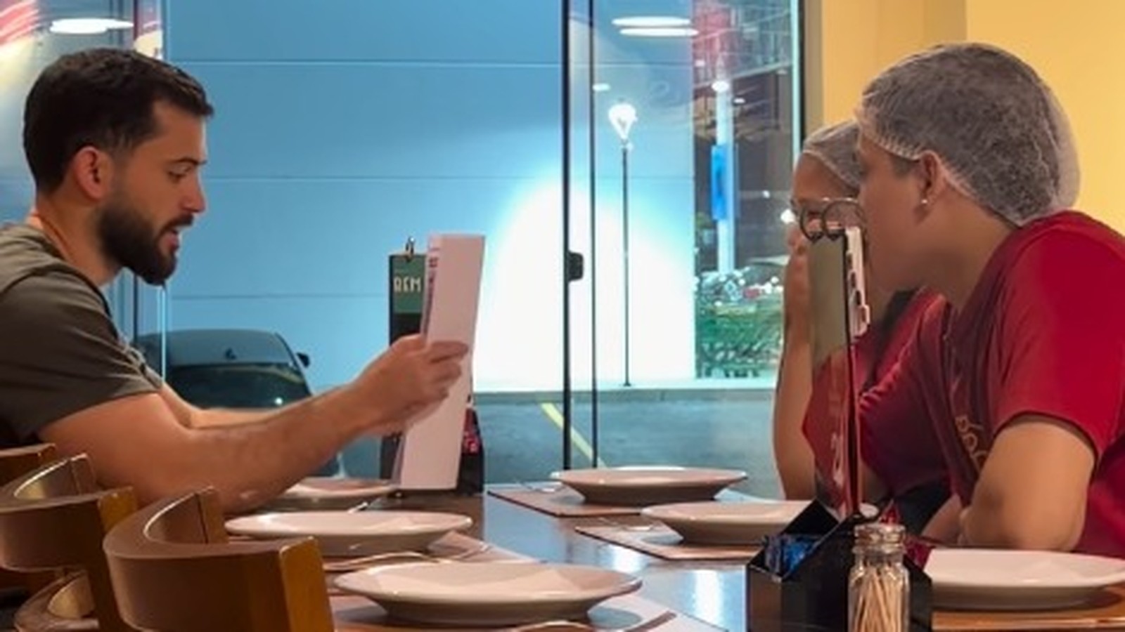 Homem com barba lendo papel para duas mulheres de touca na mesa de um restaurante