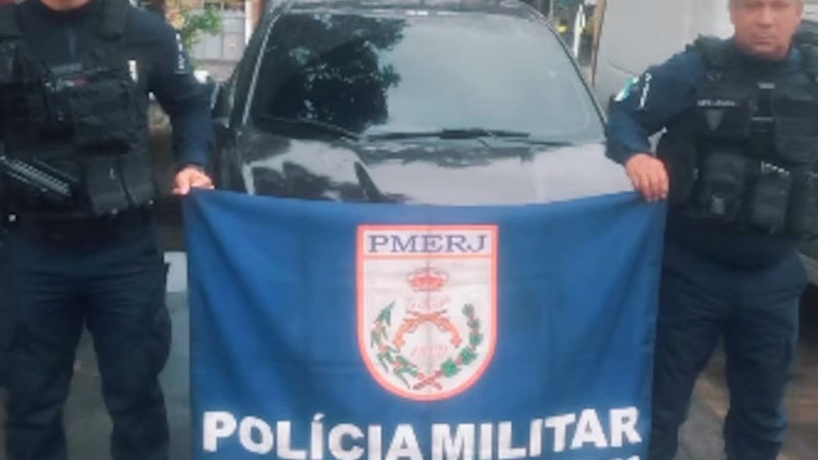 Dois policiais seguram bandeira PMERJ, Polícia Militar, em frente a carro escuro