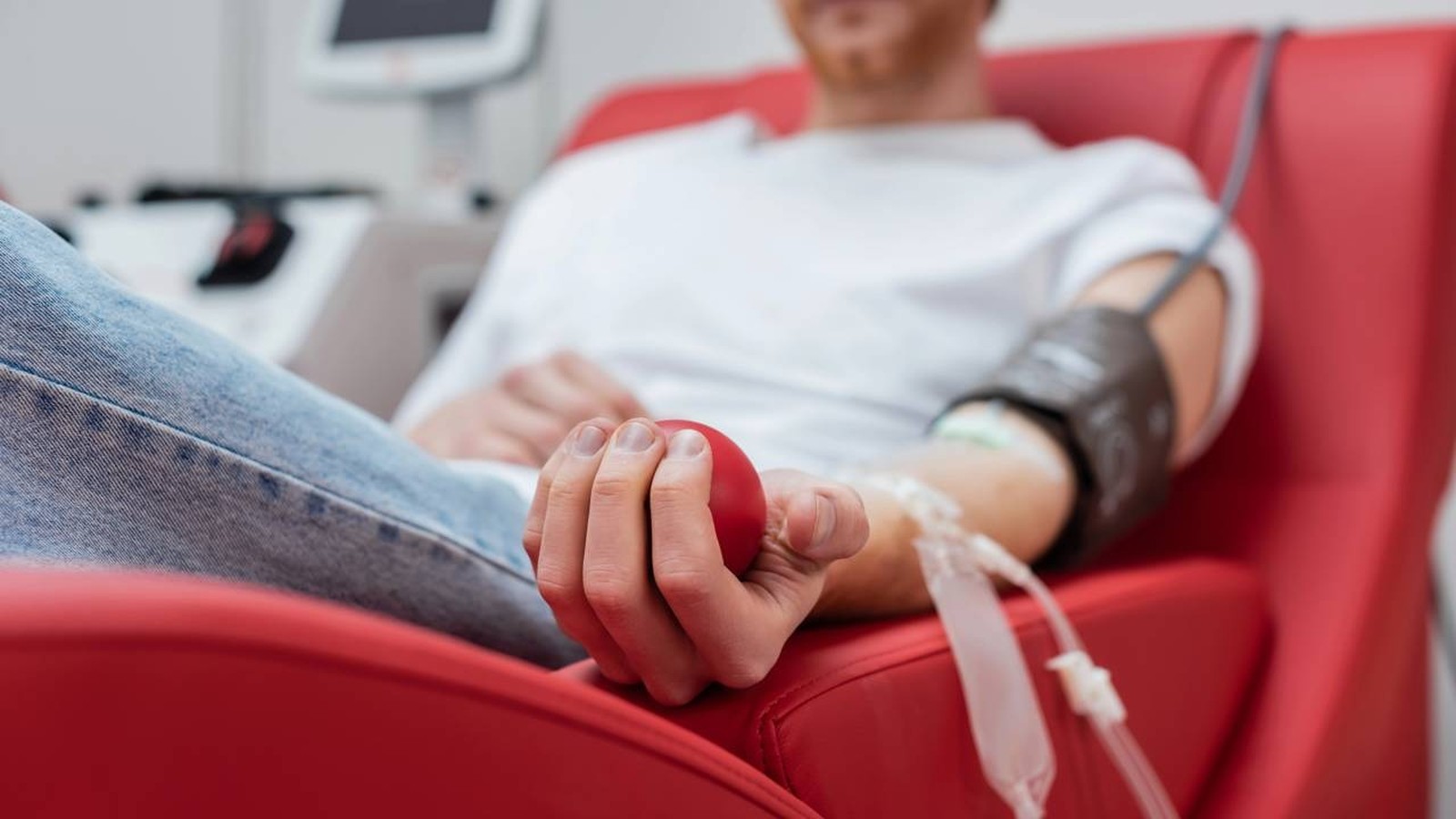 Pessoa doando sangue em cadeira vermelha, braço com equipamento, segurando bola antiestresse vermelha