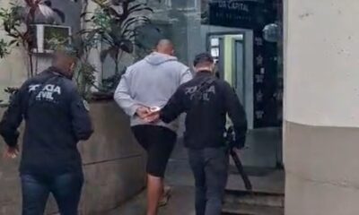 Homem algemado, moletom cinza, sendo escoltado por dois policiais civis uniformizados