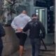 Homem algemado, moletom cinza, sendo escoltado por dois policiais civis uniformizados