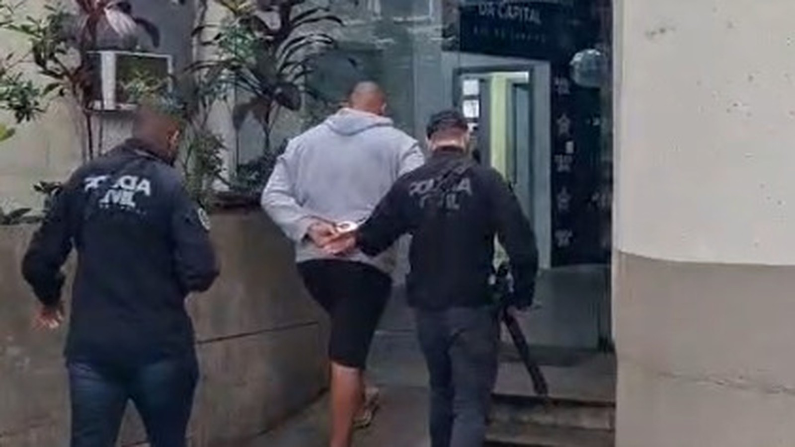 Homem algemado, moletom cinza, sendo escoltado por dois policiais civis uniformizados