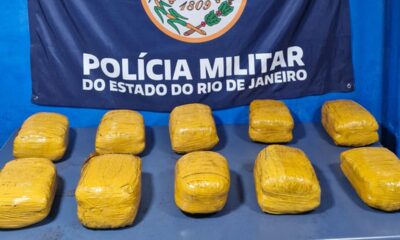Dez pacotes amarelos sobre mesa com banner da Polícia Militar do Rio de Janeiro ao fundo