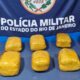 Dez pacotes amarelos sobre mesa com banner da Polícia Militar do Rio de Janeiro ao fundo
