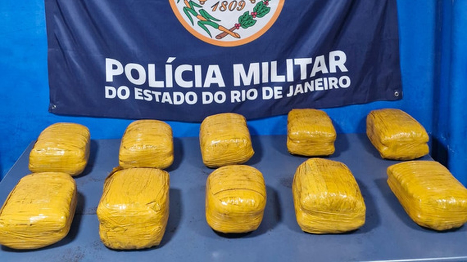 Dez pacotes amarelos sobre mesa com banner da Polícia Militar do Rio de Janeiro ao fundo