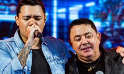 Jovem cantor com microfone e homem mais velho com colar de guitarra em palco