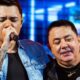 Jovem cantor com microfone e homem mais velho com colar de guitarra em palco