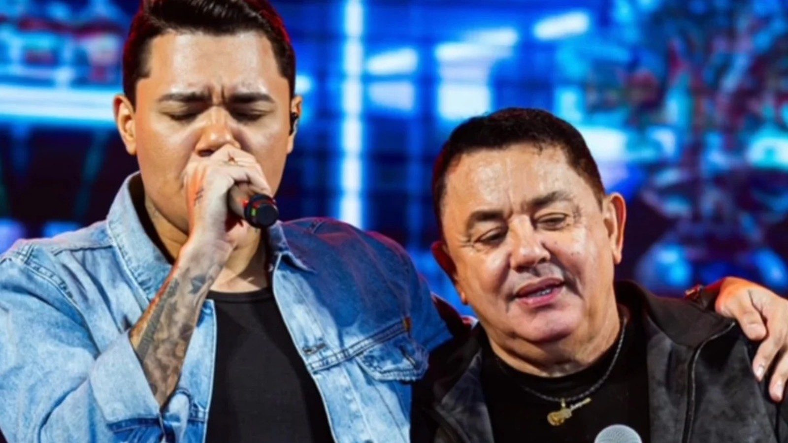 Jovem cantor com microfone e homem mais velho com colar de guitarra em palco