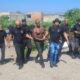 Homem musculoso, sem camisa, é escoltado por sete policiais em rua de terra com vegetação
