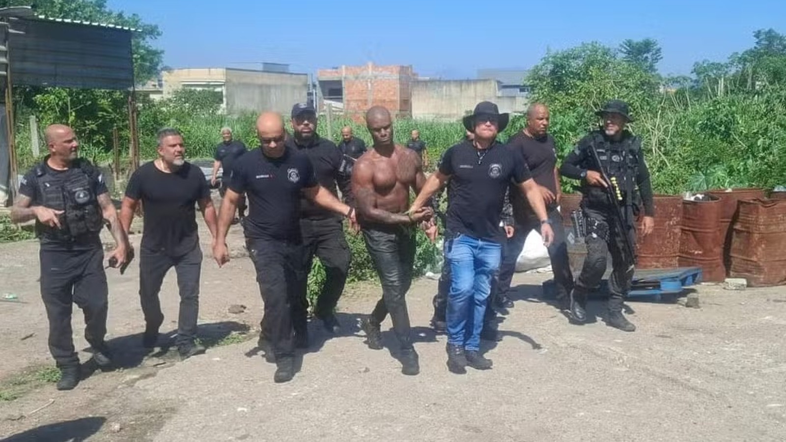 Homem musculoso, sem camisa, é escoltado por sete policiais em rua de terra com vegetação