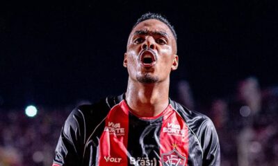 Jogador Caíque Gonçalves, suado, celebra com boca aberta, vestindo camisa preta e vermelha de patrocínios
