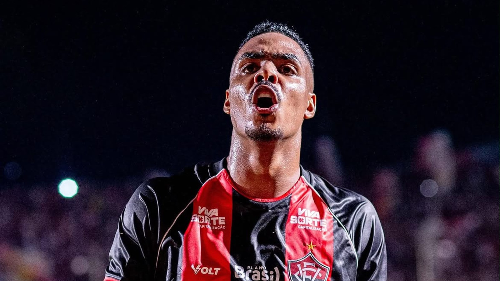 Jogador Caíque Gonçalves, suado, celebra com boca aberta, vestindo camisa preta e vermelha de patrocínios