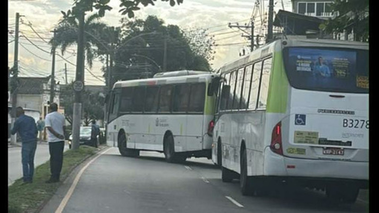 Dois ônibus brancos e verdes parados em uma rua, com homens na calçada
