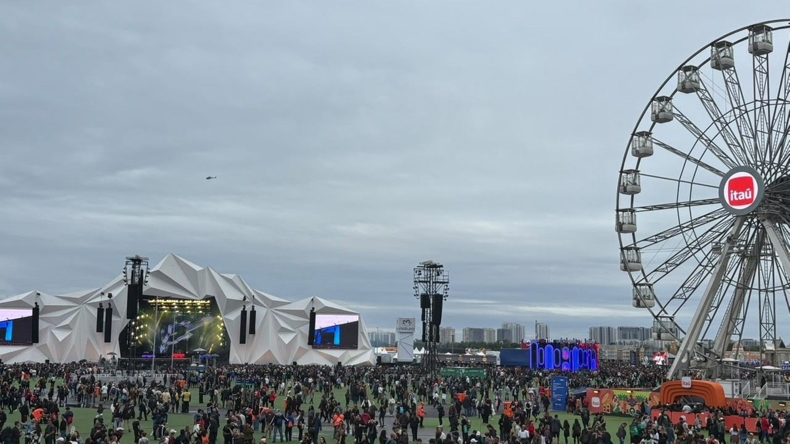 Multidão em festival com palco grande, roda-gigante Itaú e helicóptero no céu nublado