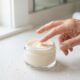 Pote de hidratante facial aberto com creme visível e mão de mulher madura tocando o produto.