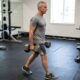 Pessoa com mais de 50 anos caminhando com dois halteres ao lado do corpo e mantendo a postura ereta.