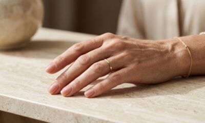Mão feminina madura com unhas curtas esmaltadas em nude rosado brilhante sobre superfície clara.