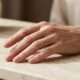 Mão feminina madura com unhas curtas esmaltadas em nude rosado brilhante sobre superfície clara.