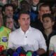 Jair Bolsonaro discursa em pódio com microfones; homens de camisas do Brasil atrás