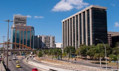 Paisagem urbana com prédios altos, rodovia movimentada, carros, ponte e palmeiras sob céu azul