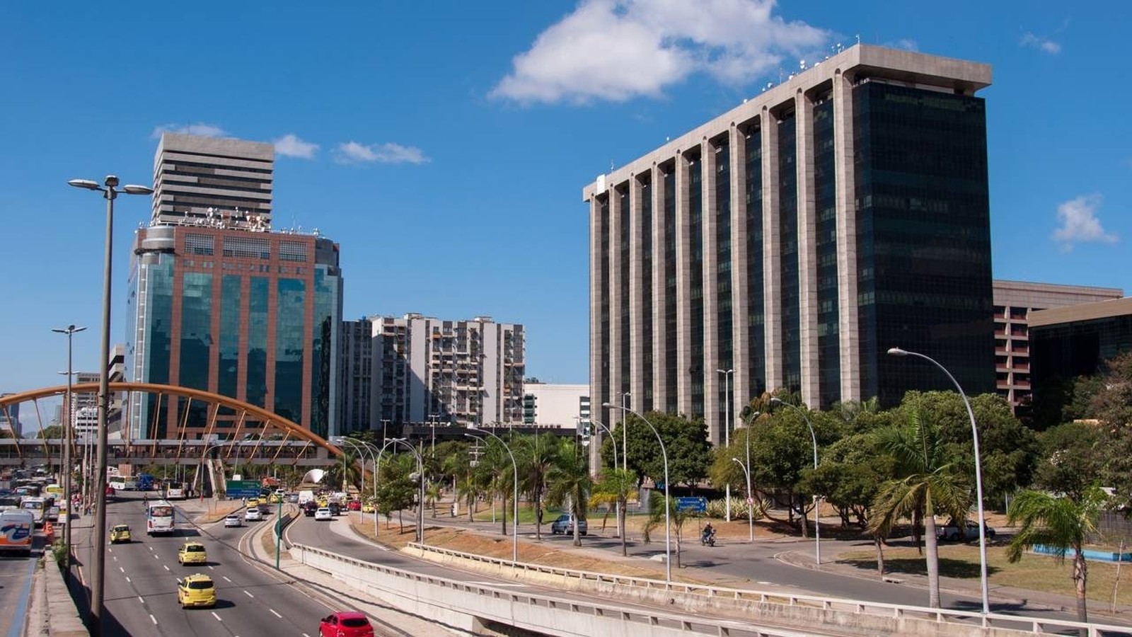 Paisagem urbana com prédios altos, rodovia movimentada, carros, ponte e palmeiras sob céu azul