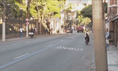 Rua da cidade com faixa "ÔNIBUS" no asfalto, carros, moto e pedestres na calçada