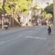 Rua da cidade com faixa "ÔNIBUS" no asfalto, carros, moto e pedestres na calçada
