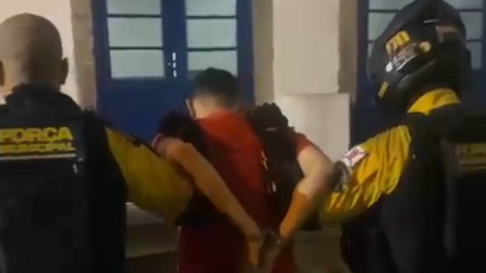 Dois agentes da Força Municipal escoltam homem algemado em camisa vermelha