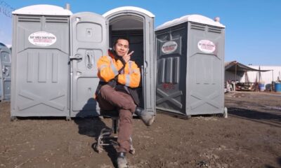 Daniel Tom entre dois banheiros químicos cinzas da Bay Area Sanitation, fazendo sinal de paz
