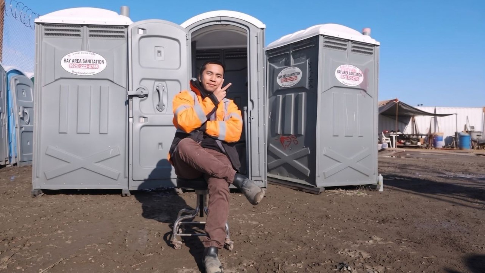 Daniel Tom entre dois banheiros químicos cinzas da Bay Area Sanitation, fazendo sinal de paz