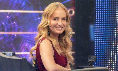 Angélica, cabelos loiros, sorri em estúdio de TV usando blusa vermelha e microfone de lapela