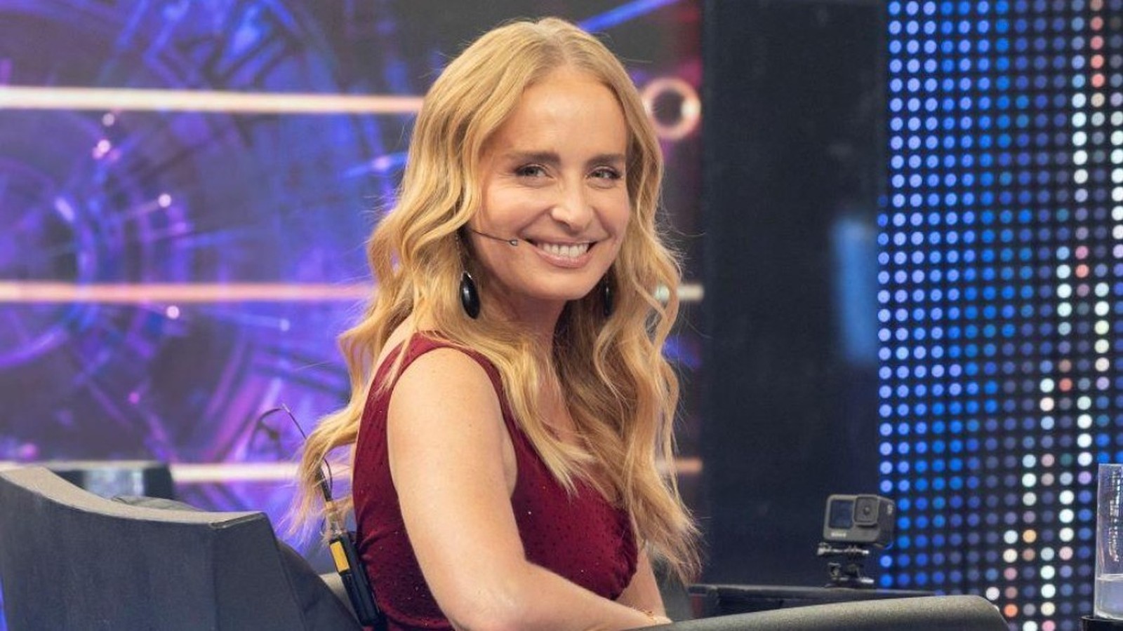 Angélica, cabelos loiros, sorri em estúdio de TV usando blusa vermelha e microfone de lapela