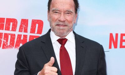 Arnold Schwarzenegger sorrindo, de terno e gravata vermelha, fazendo sinal de positivo