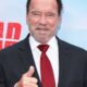 Arnold Schwarzenegger sorrindo, de terno e gravata vermelha, fazendo sinal de positivo