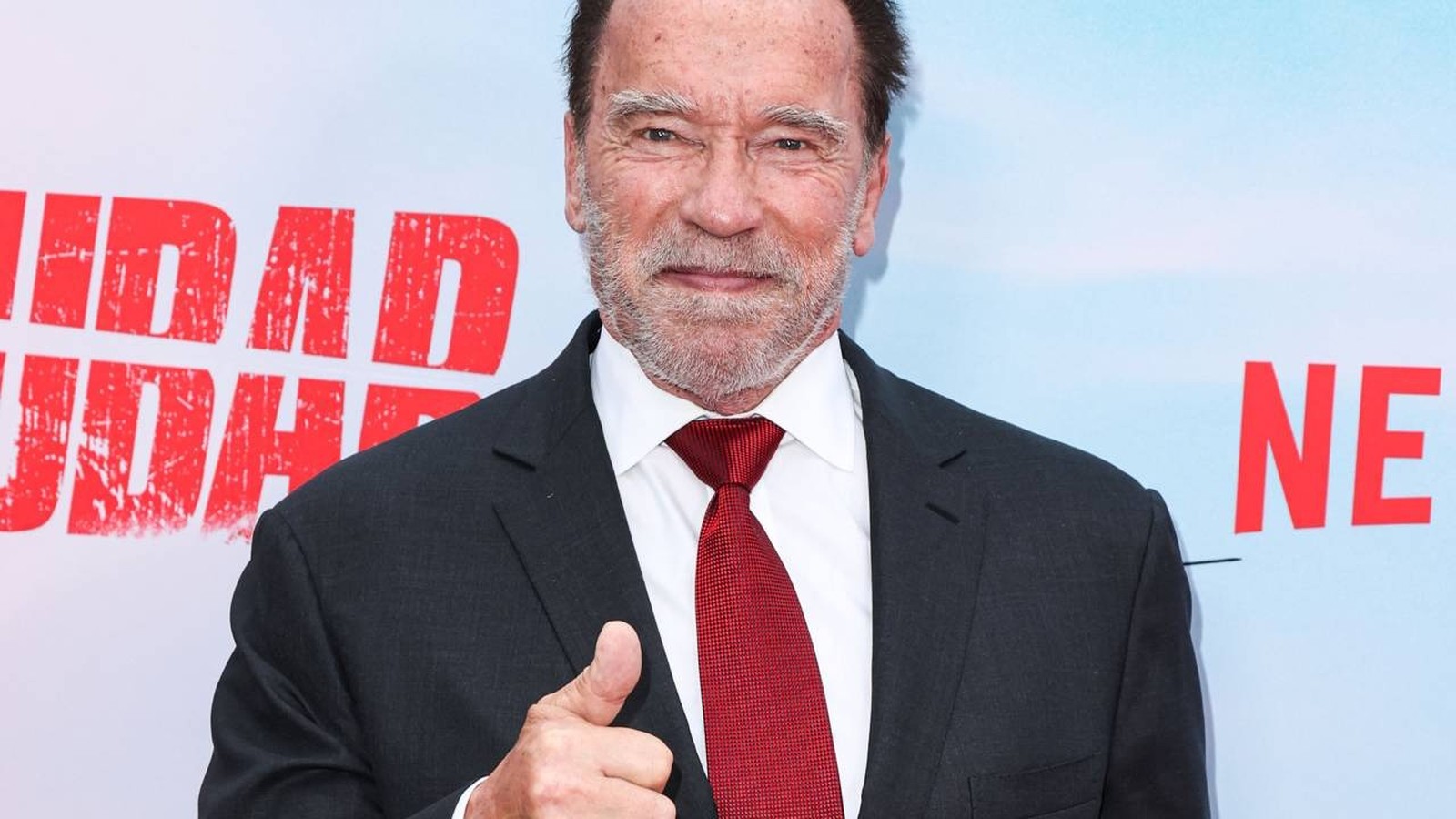 Arnold Schwarzenegger sorrindo, de terno e gravata vermelha, fazendo sinal de positivo