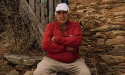 Homem idoso de boné branco e camisa vermelha sentado com braços cruzados, em frente a parede de pedra