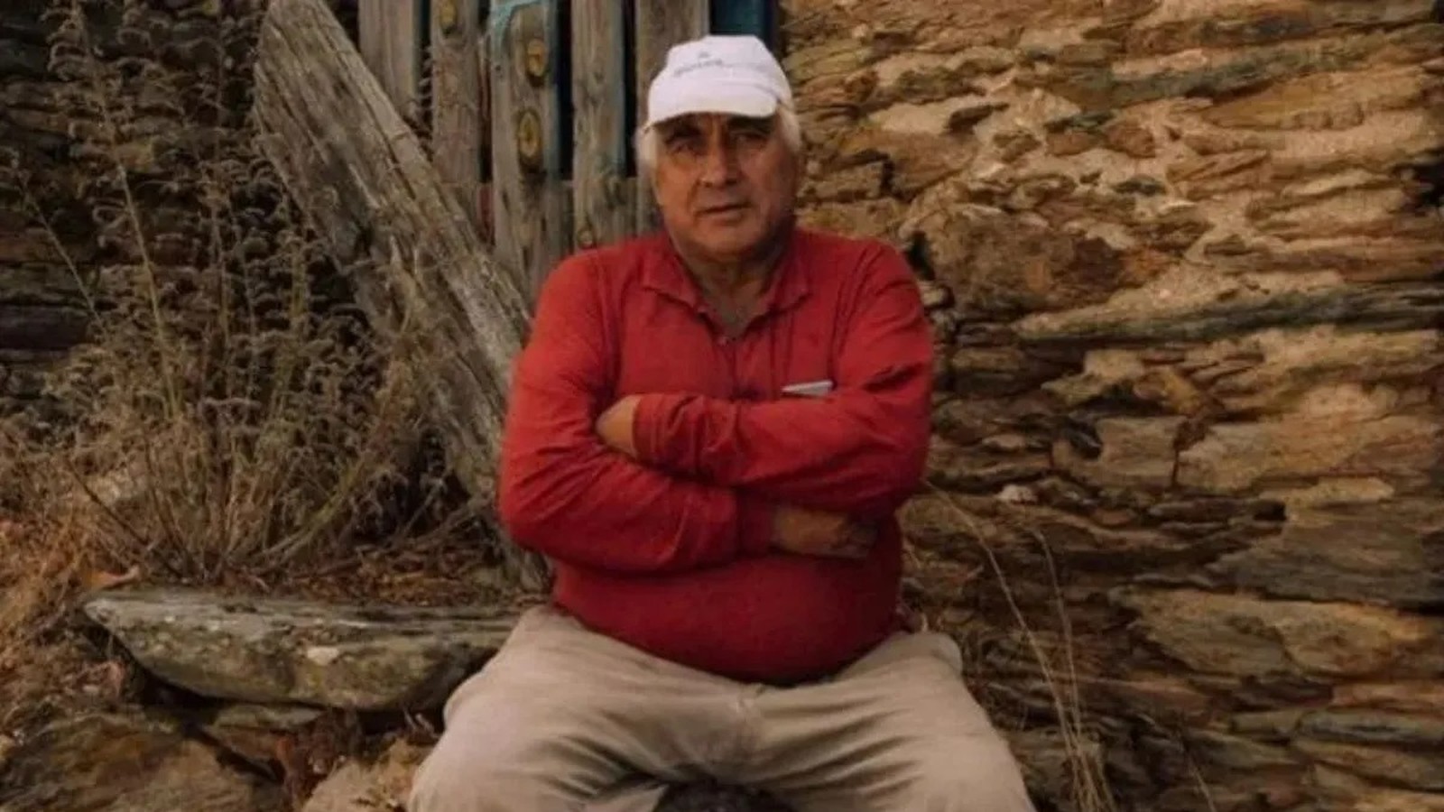 Homem idoso de boné branco e camisa vermelha sentado com braços cruzados, em frente a parede de pedra
