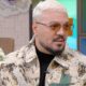 Homem de cabelo loiro platinado, barba, óculos de sol e jaqueta estampada olhando à direita
