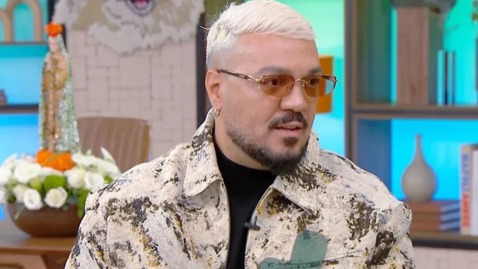 Homem de cabelo loiro platinado, barba, óculos de sol e jaqueta estampada olhando à direita