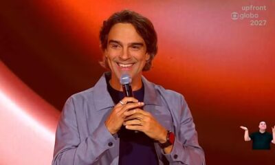 Tadeu Schmidt sorri ao microfone em palco laranja, texto upfront globo 2027, intérprete de libras