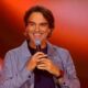 Tadeu Schmidt sorri ao microfone em palco laranja, texto upfront globo 2027, intérprete de libras