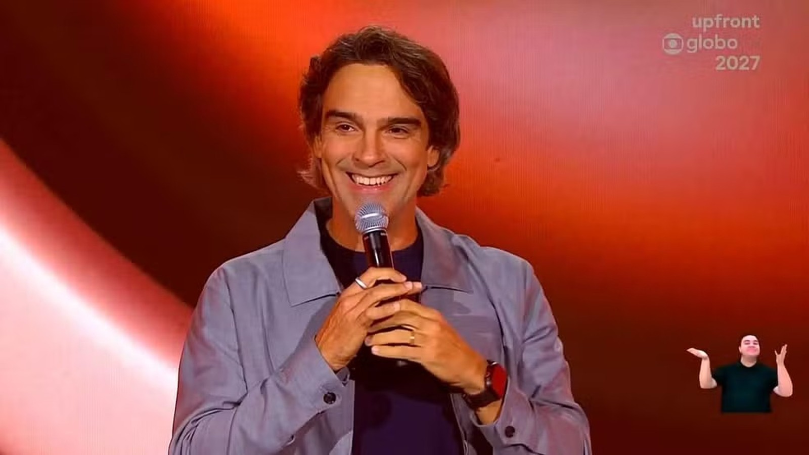 Tadeu Schmidt sorri ao microfone em palco laranja, texto upfront globo 2027, intérprete de libras