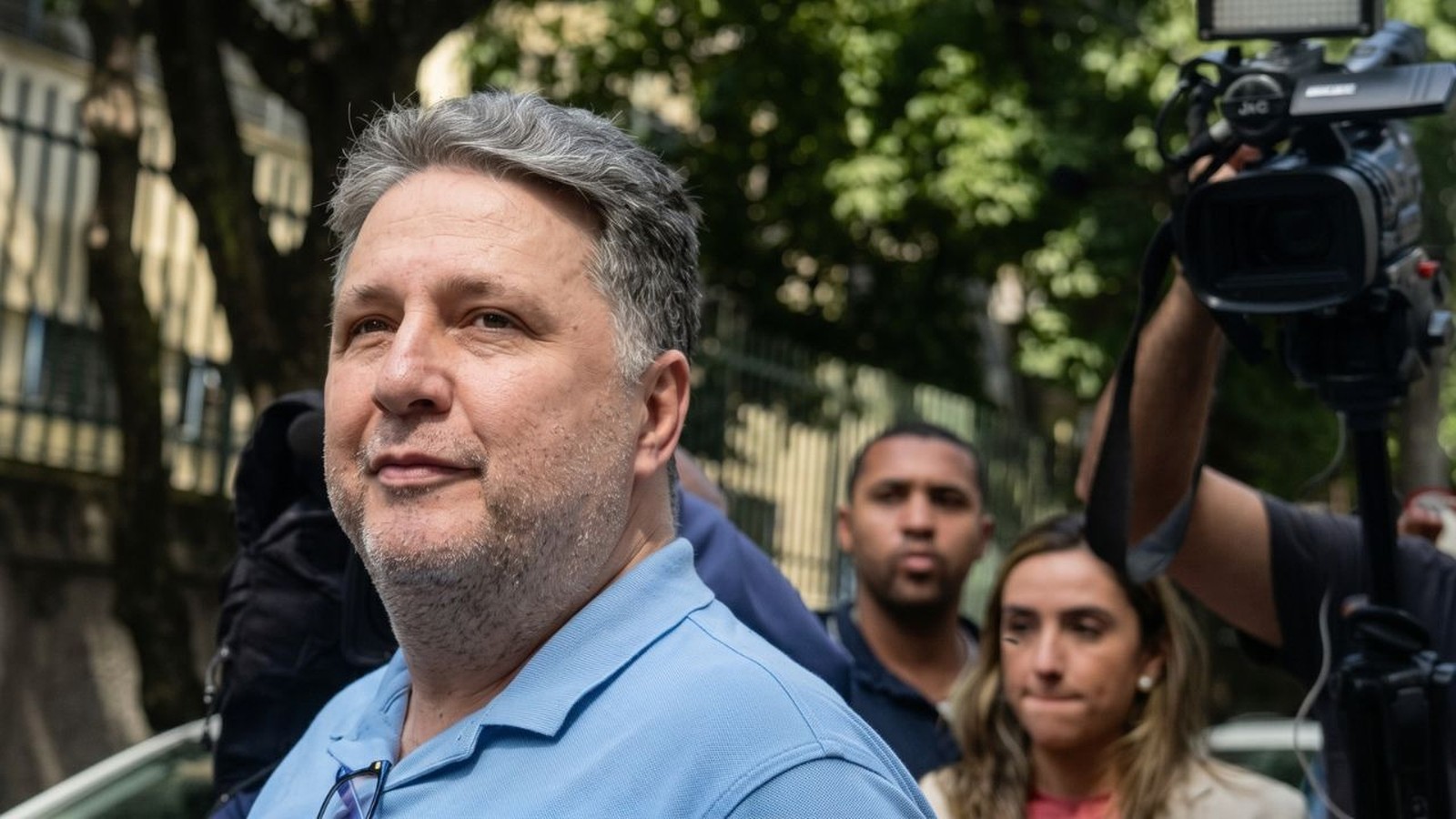 Homem com cabelo grisalho, camisa polo azul, óculos no colarinho, em meio a equipe de filmagem